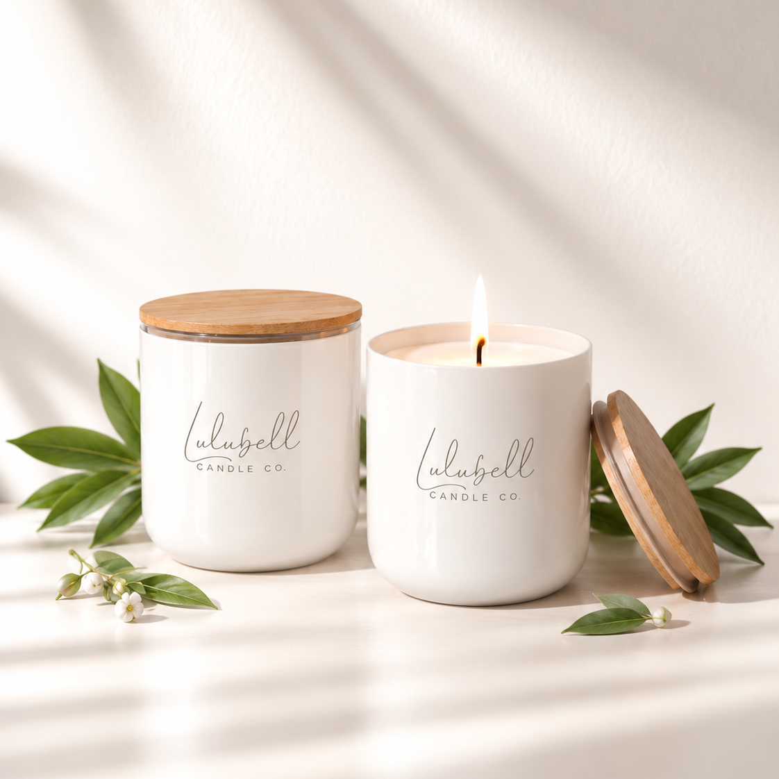 Two white scented candles from Lulubell Candle Co. on a white surface with soft leaf shadows.