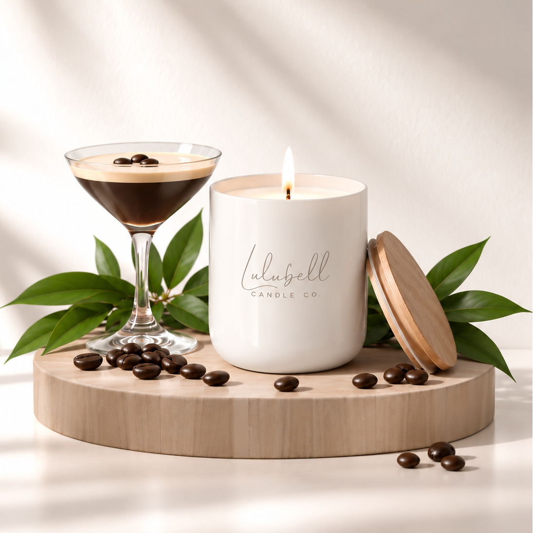 A white scented candle labeled 'Lulubell Candle Co' sits on a light wood tray beside a martini glass filled with espresso and coffee beans, with green leaves and a wooden lid leaning against it in a softly lit setting.