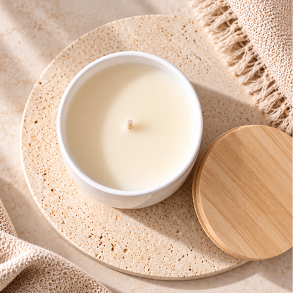A white ceramic candle jar with a single wick and cream-colored wax, shown on a round speckled beige stone coaster with a light wood lid beside it and soft beige textiles in the background.
